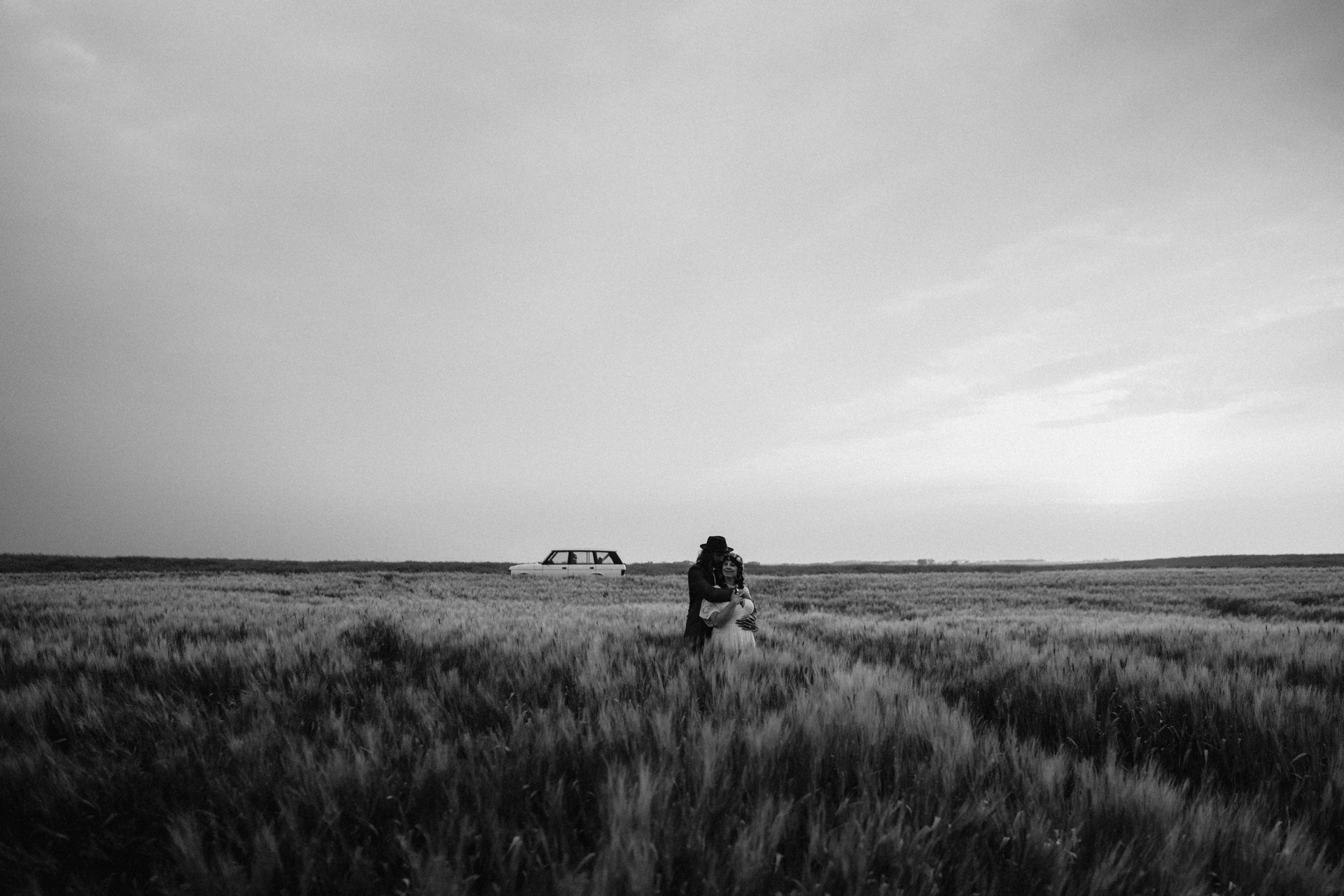 Cayle et Nathaniel enlacés dans un immense champ de blé sous un ciel pâle, une vieille familiale stationnée à l’horizon